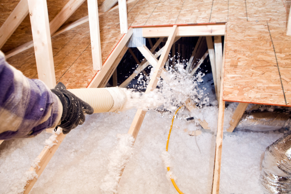 A person sprays loose-fill insulation into an attic, covering the floor between wooden beams with white insulating material.