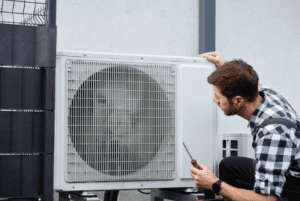 A man inspects an outdoor air conditioning unit with a screwdriver in hand, appearing to perform maintenance or repairs.