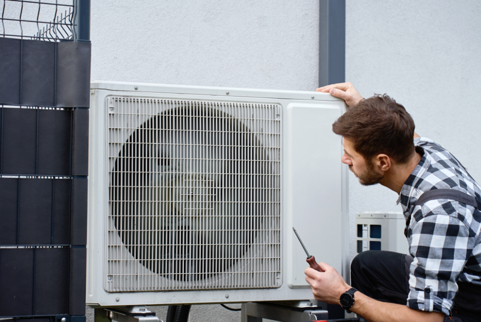 A man inspects an outdoor air conditioning unit with a screwdriver in hand, appearing to perform maintenance or repairs.