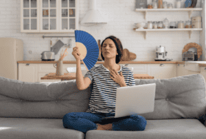 A woman sits on a sofa with a laptop on her lap, fanning herself and looking hot, in a bright kitchen setting.
