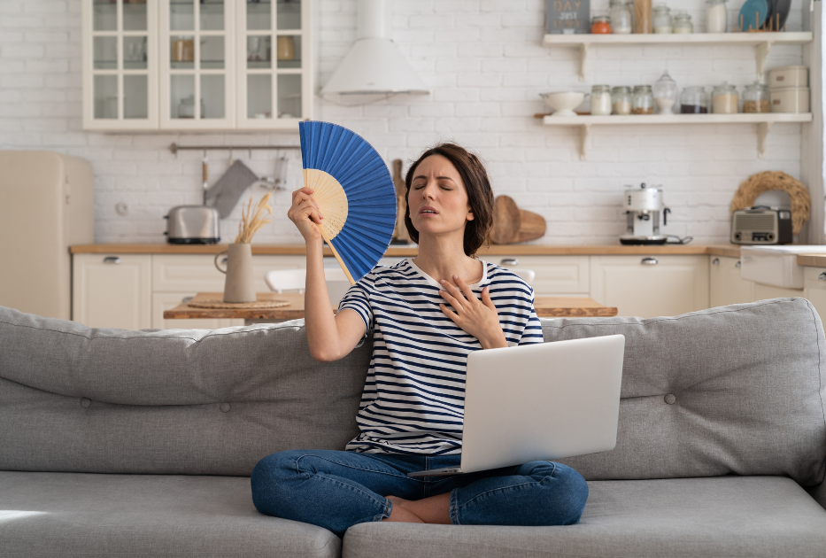 A woman sits on a sofa with a laptop on her lap, fanning herself and looking hot, in a bright kitchen setting.