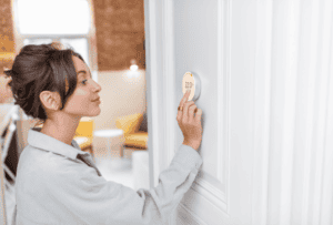 Woman adjusting a digital thermostat on a white wall, setting the temperature to 22°C in a modern, brightly lit room.
