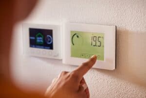 A person’s finger presses a button on a digital thermostat, which displays a temperature reading of 19.5 degrees Celsius. Another control panel is mounted nearby on the wall.