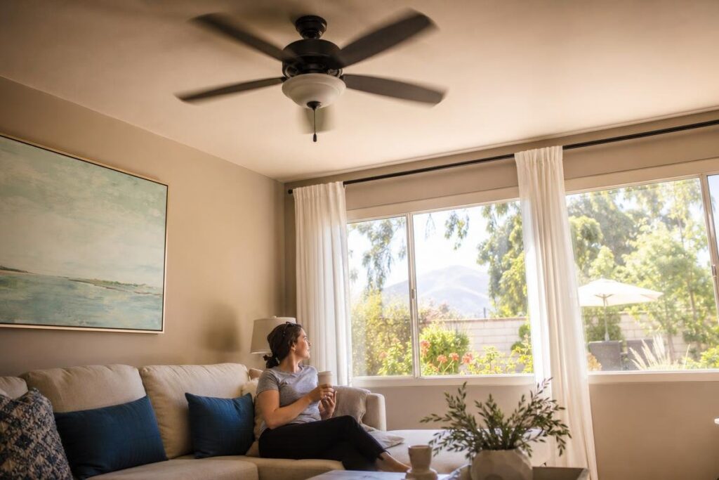 Woman sits on a beige couch holding a mug, looking out a large window at a sunny yard. A ceiling fan and wall art are visible in the living room.