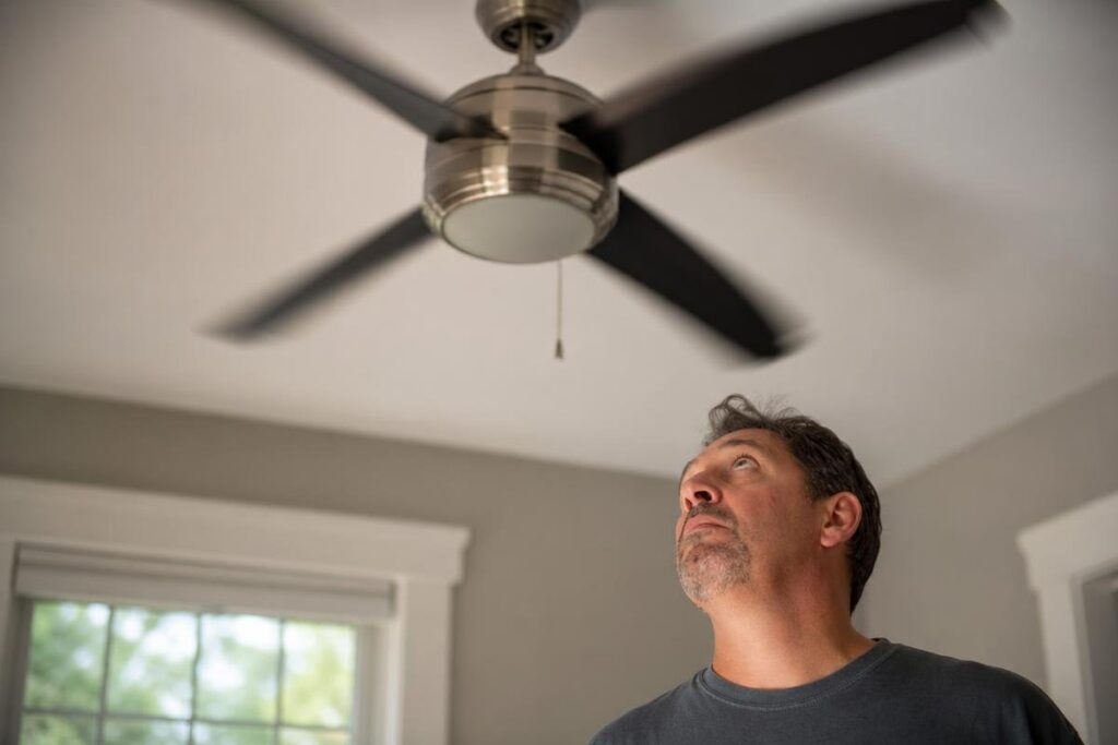 A man stands indoors and looks up at a ceiling fan with black blades that is turned on. Natural light enters through a window in the background.