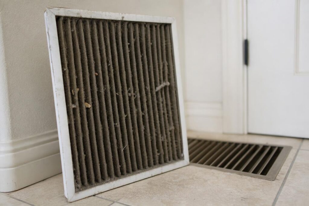 A dirty air filter leaning against a wall next to a floor vent on a tiled surface.