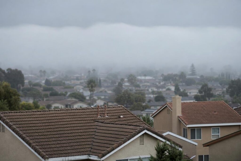 Rows of suburban houses with tile roofs sit under a cloudy, foggy sky that partially obscures the distant landscape.